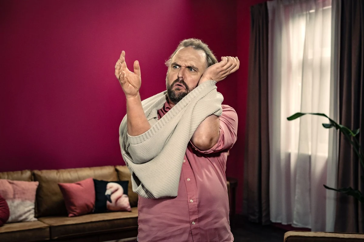 Ronny Schuster in pinkfarbenem Hemd beim Ausziehen eines hellen Pullovers.