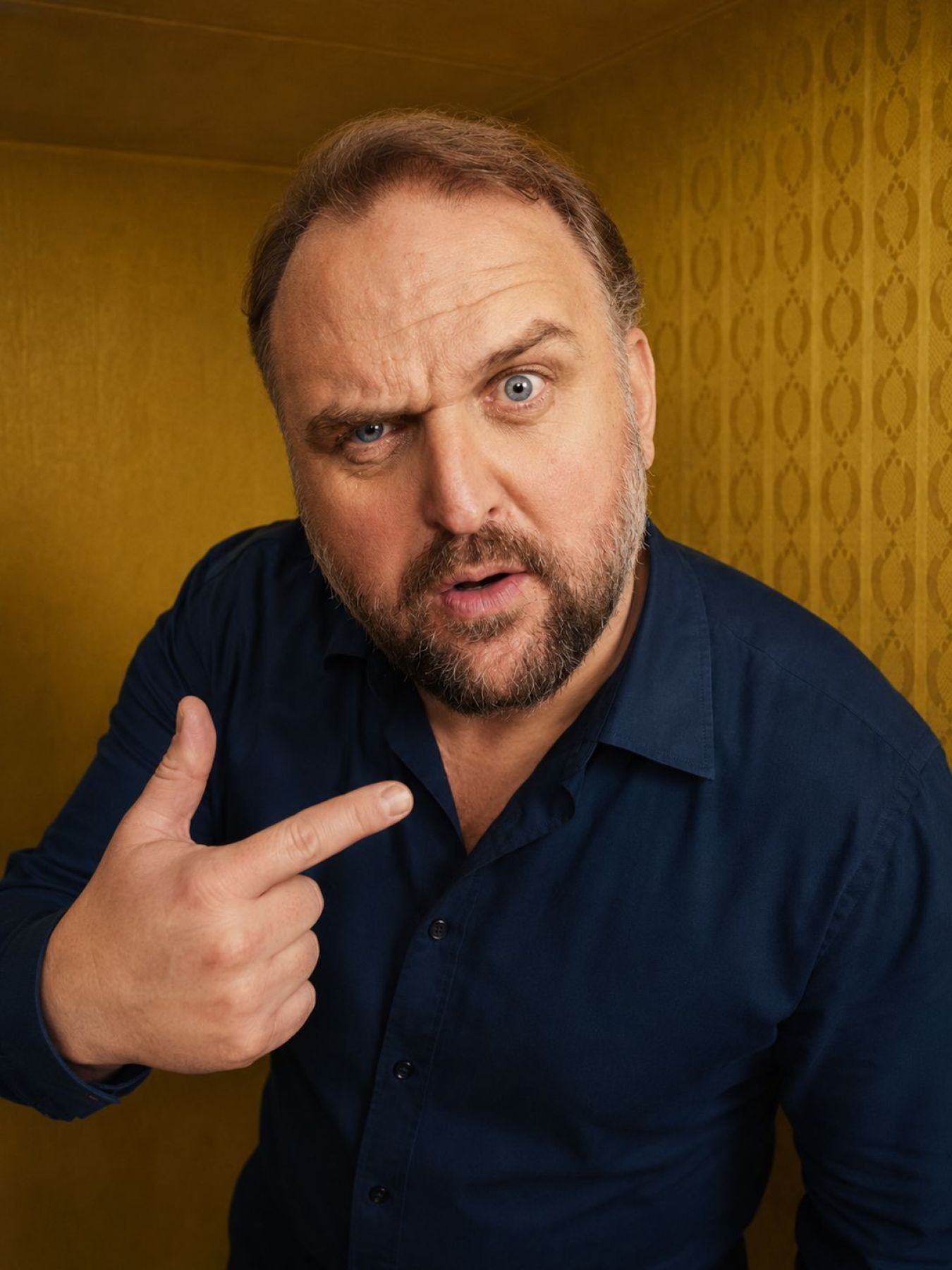 Ronny Schuster im dunkelblauen Hemd mit fragendem Blick und ausgestreckten Fingern.