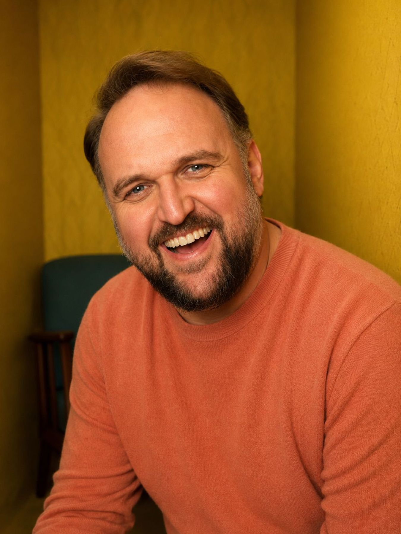 Ronny Schuster lächelt im orangefarbenen Pullover vor gelber Wand.