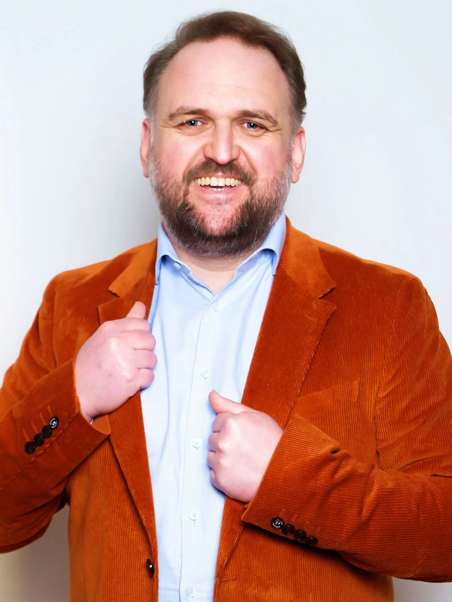 Ronny Schuster lächelt in orangefarbenem Sakko und hellblauem Hemd.