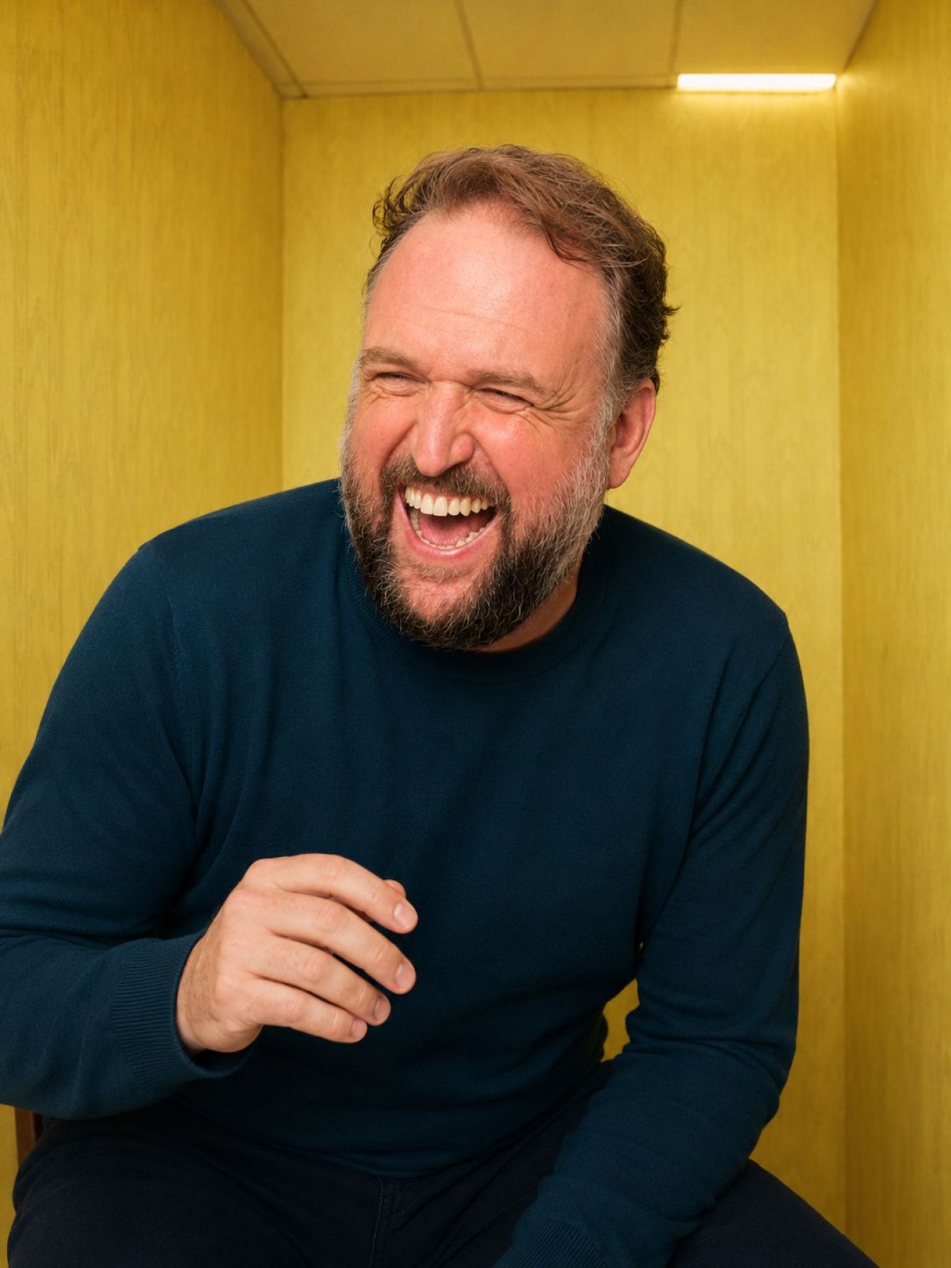Ronny Schuster lacht in einem dunkelblauen Pullover vor gelber Wand.